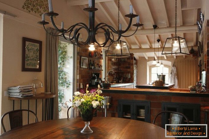 La salle à manger et la cuisine sont décorées dans un style campagnard. Ce qui est remarquable est un lustre au-dessus de la table, qui illumine l'espace à l'aide de bougies en cire ordinaires. Idée de design mince, car dans la pièce il y a aussi un éclairage traditionnel, fonctionnant à partir du réseau électrique.
